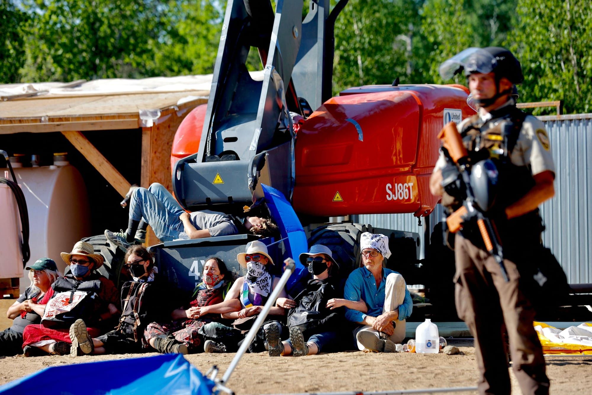 climate activists sit on the ground in front of construction equipment. a police officer in riot gear stands in the foregroun