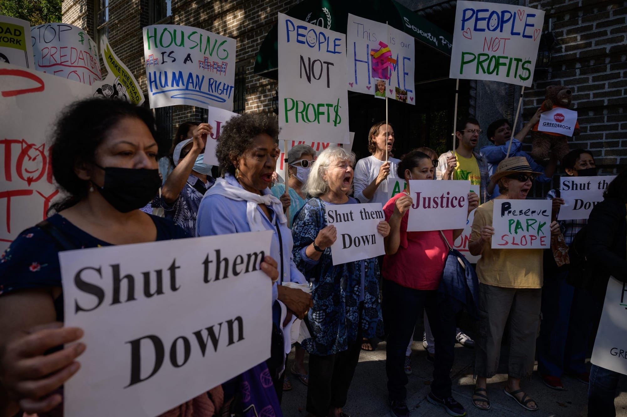 a photograph of a crowd of protesters facing to the right of the camera. they hold signs saying "shut them down" and "people