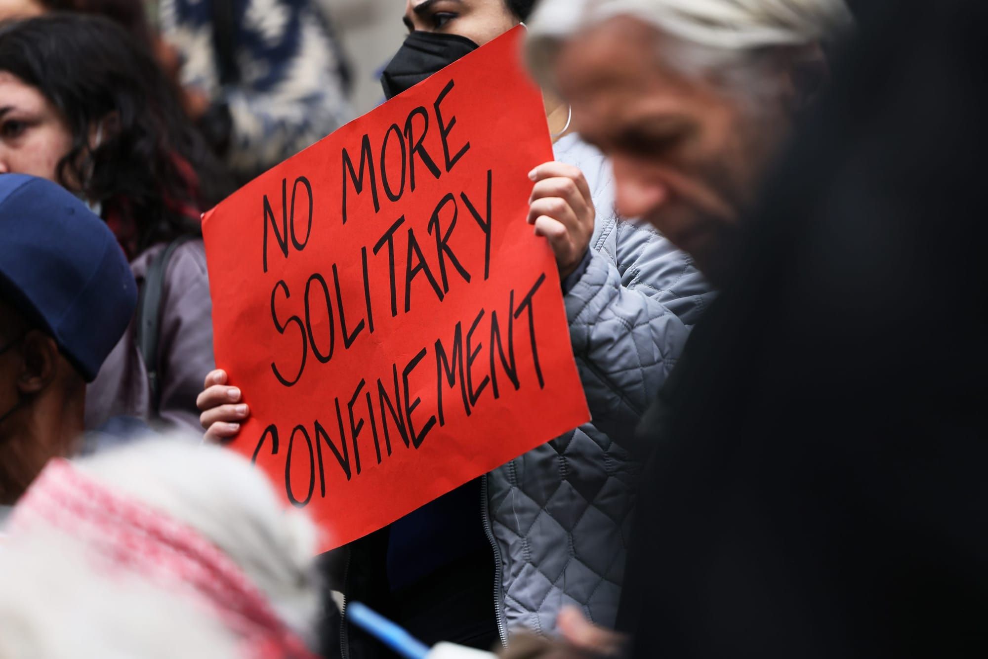 color photograph of a person in a crowd of protesters holding a red paper poster with black ink text reading "no more solitar