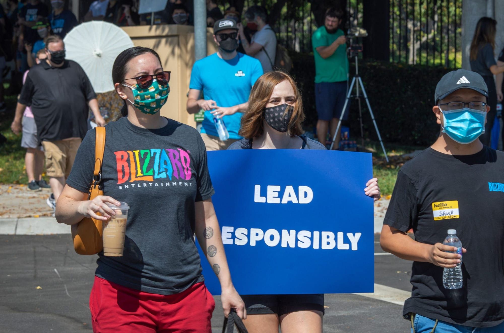 color photograph of an outdoor protest of video game workers. one is wearing a grey t-shirt with a rainbow-colored logo for t