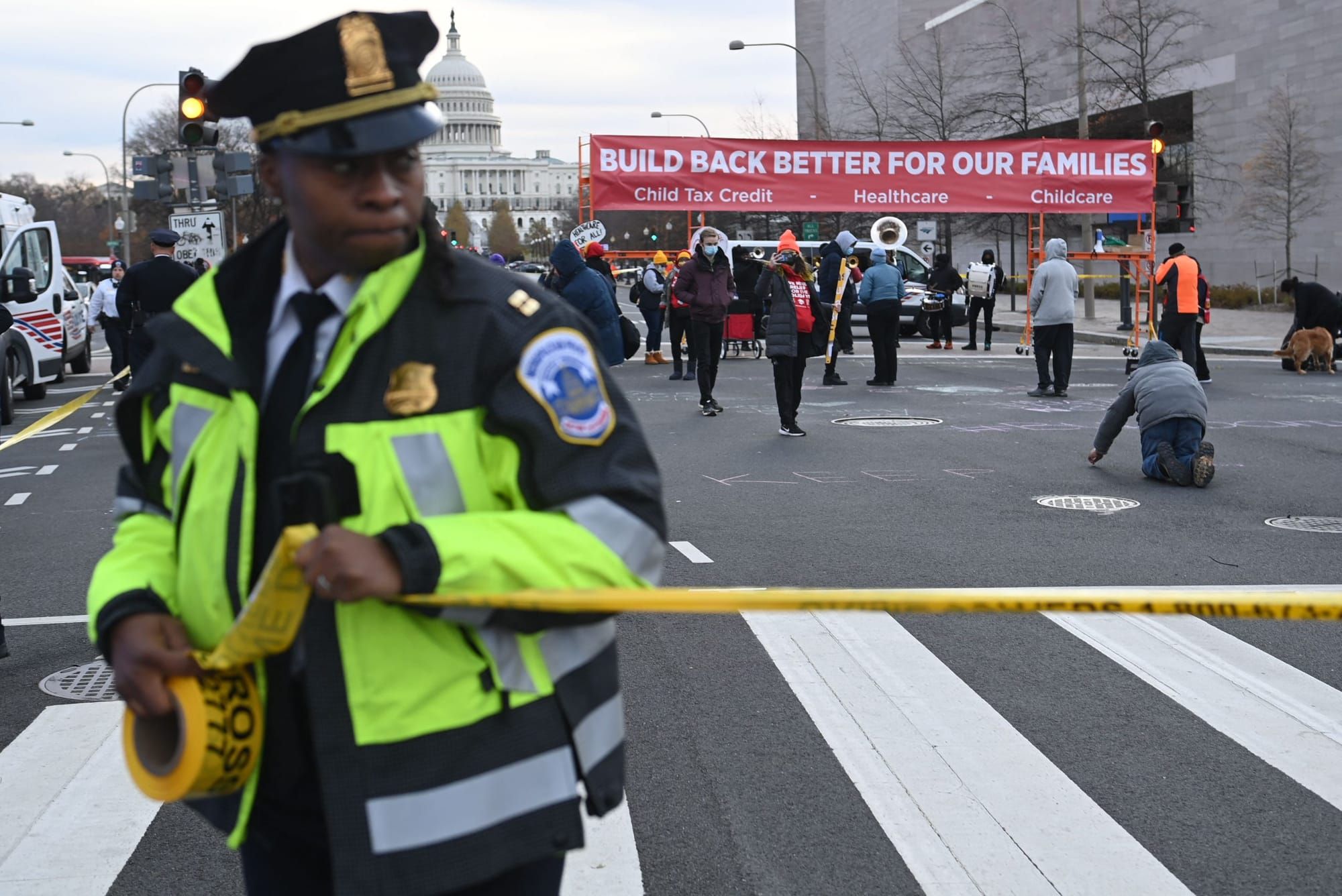 color photograph of a police officer in a yellow safety vest pulling a line of yellow caution tape across the foreground. in