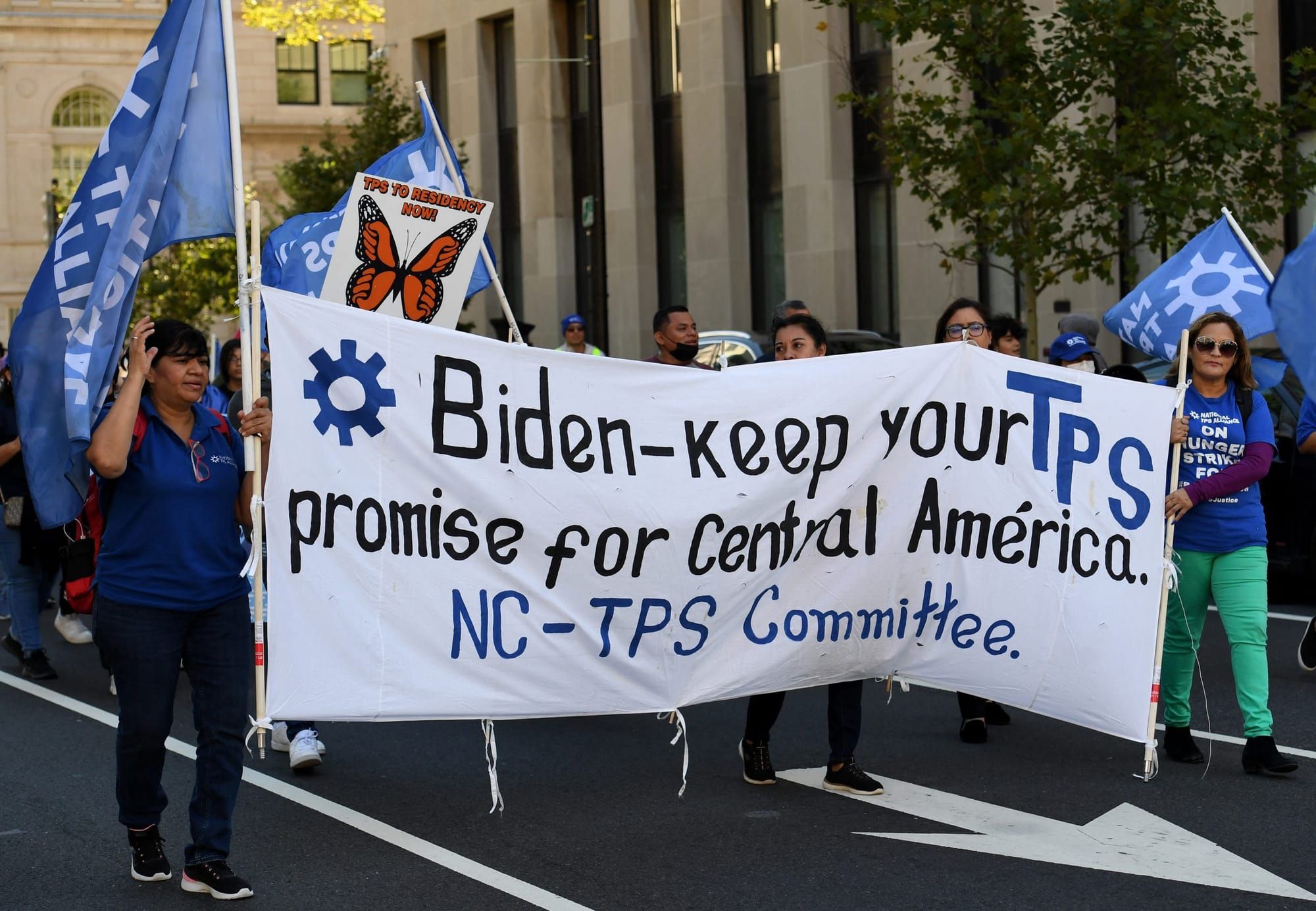 protesters hold a large white banner that spans several meters. The banner has black text that reads "Biden—keep your TPS pro