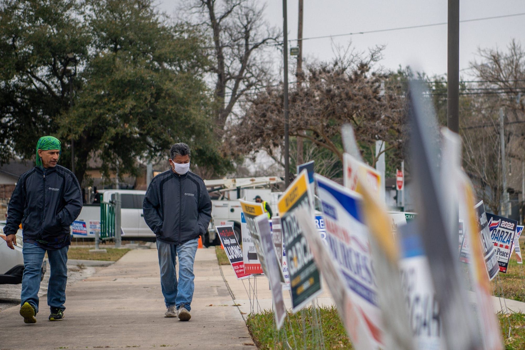 color photograph of two men wearing jeans, winter jackets, and masks walking on a sidewalk next to a row of political campaig