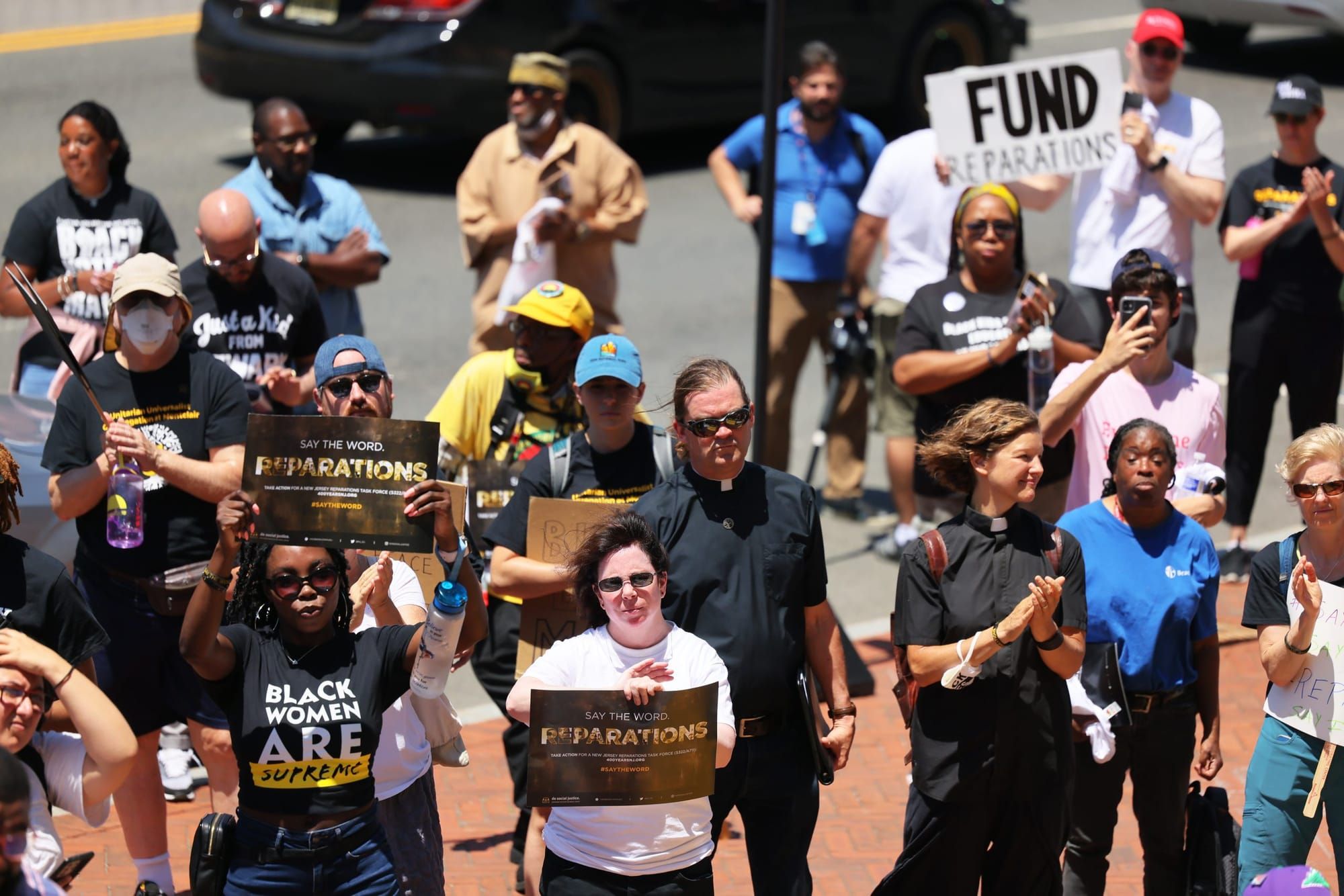 color photograph of an outdoor protest where a diverse crowd holds signs calling for reparations