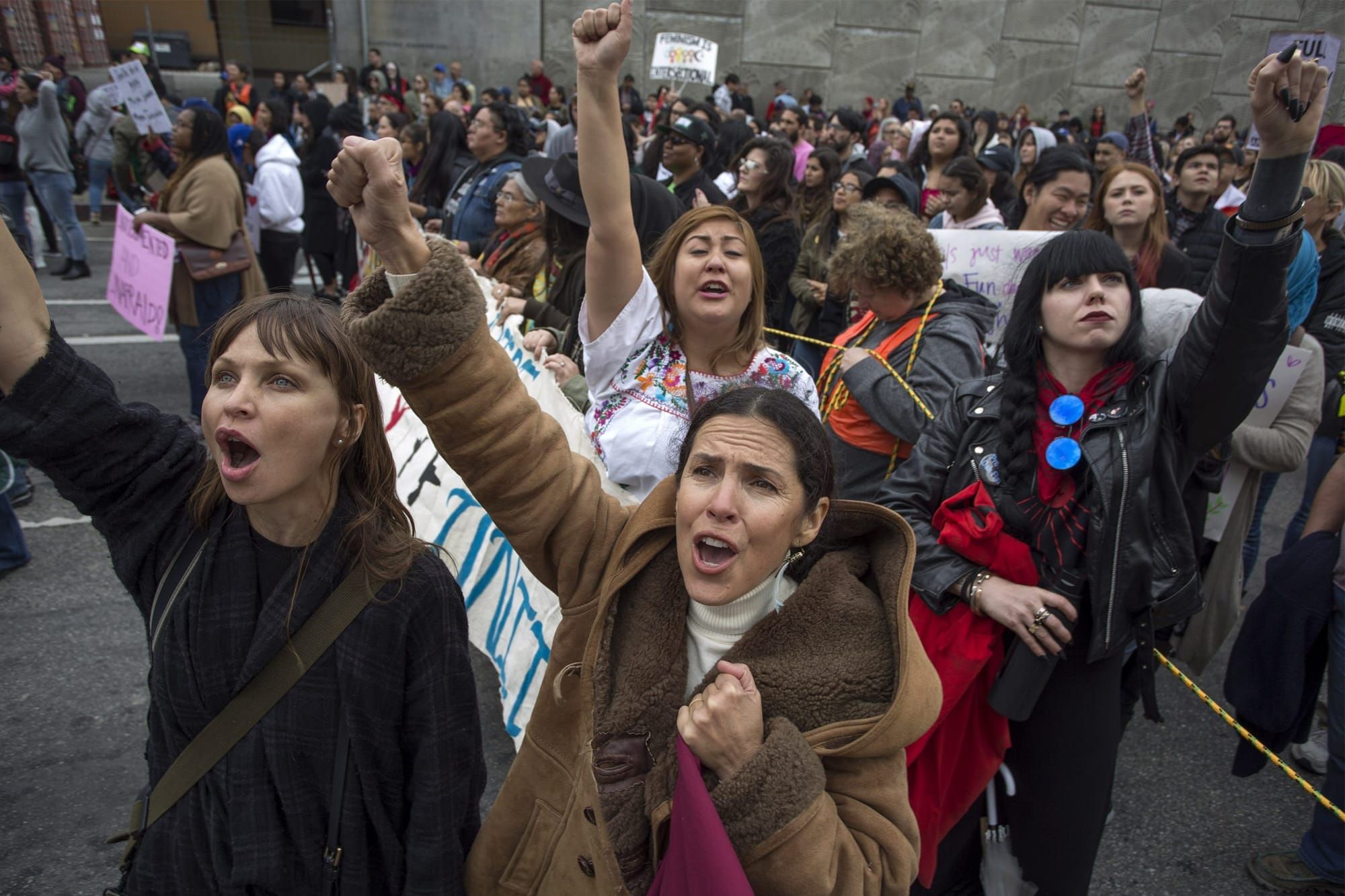 color photograph of a crowd of women standing outside wearing winter coats. they stand facing the camera with their fists rai