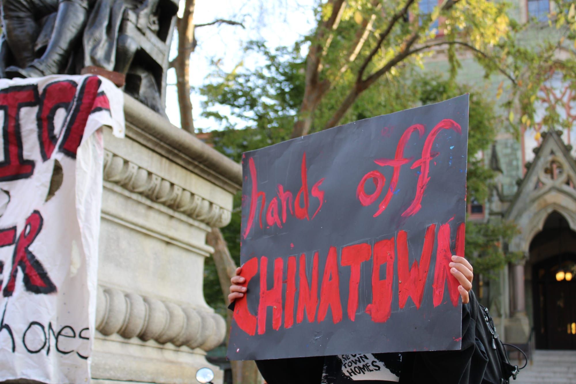 color photograph of a person standing outside holding a black paper poster with red text reading "hands off chinatown" obscur