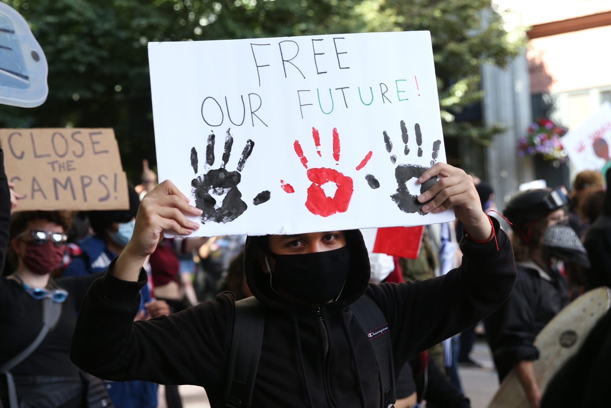 color photograph of an outdoor protest, mid-close-up of a protester wearing all black while holding up a white paper sign tha