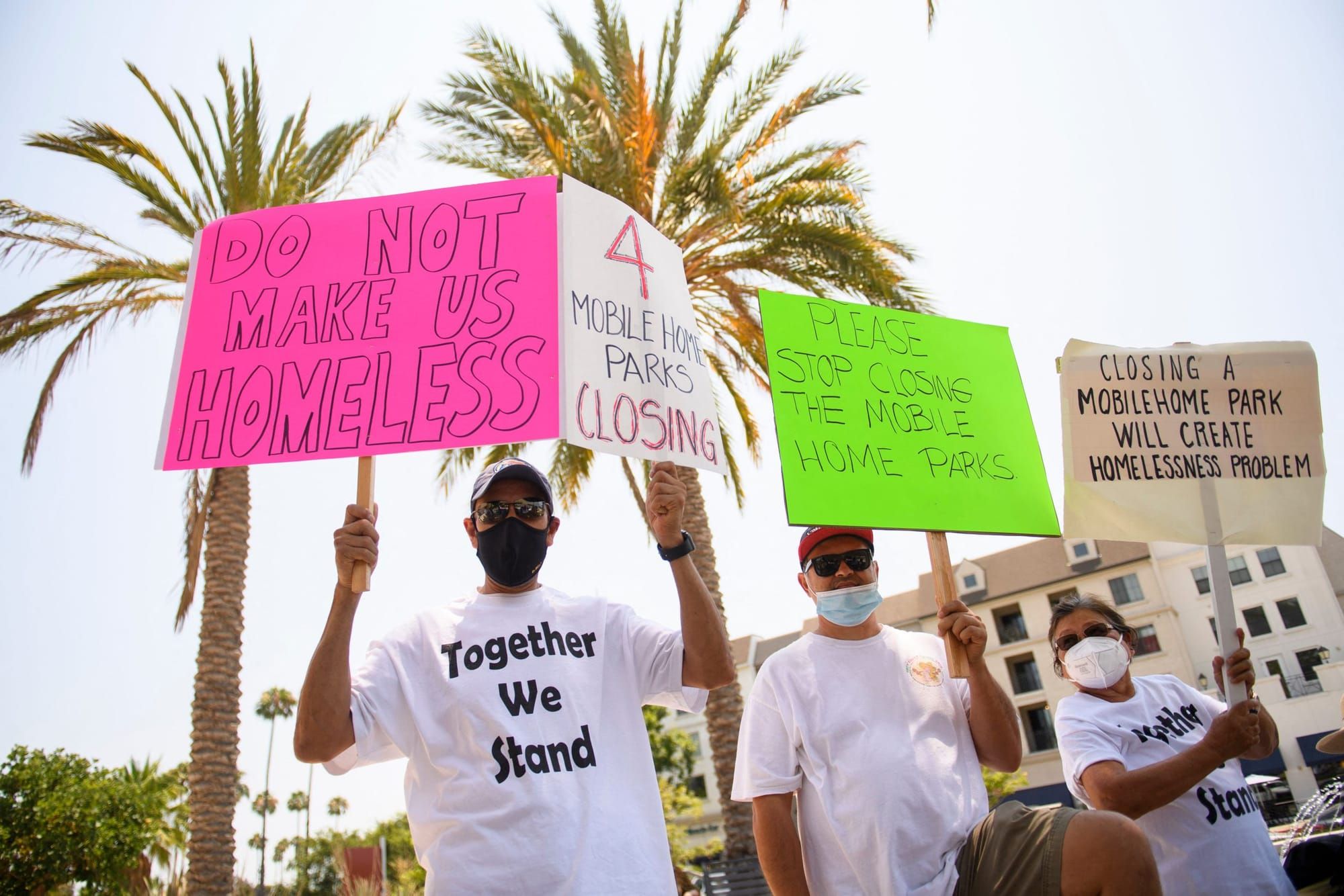 color photograph of an outdoor protest. people wear white t-shirts and hold up pink, white, and green paper signs on wooden p