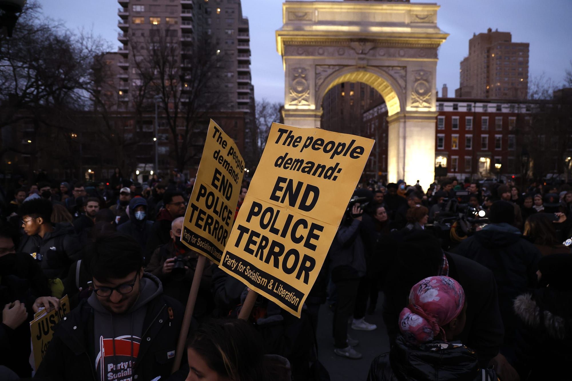 color photograph of an outdoor protest in the early evening. people in the foreground are not lit, but a decorative stone arc
