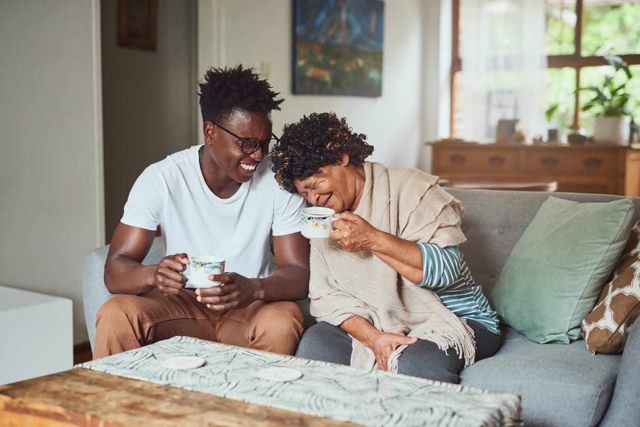 color photograph stock image of a young black man sitting on a grey sofa with his mother. they are both smiling and holding c