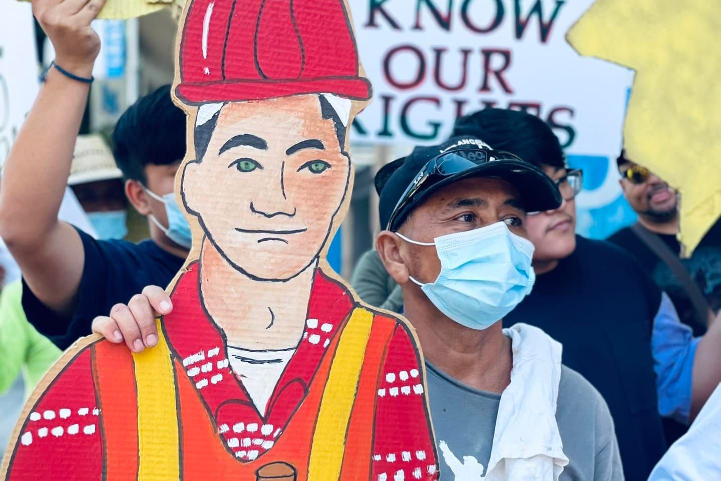 color photograph of a masked person standing next to a cardboard cutout of a worker, with a sign in the background that reads
