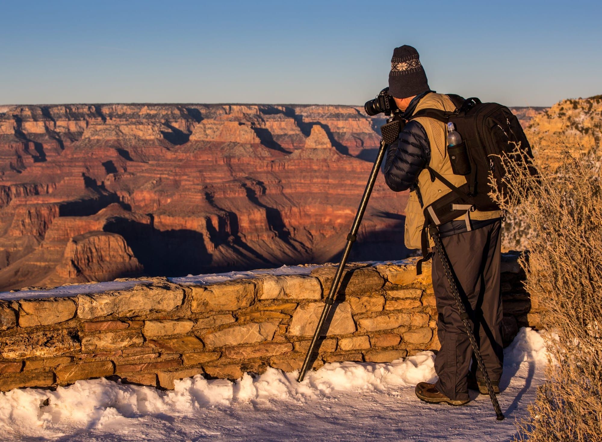 color photograph of a person wearing a hat, brown jacket, and dark brown long pants leaning towards a camera on a tripod over