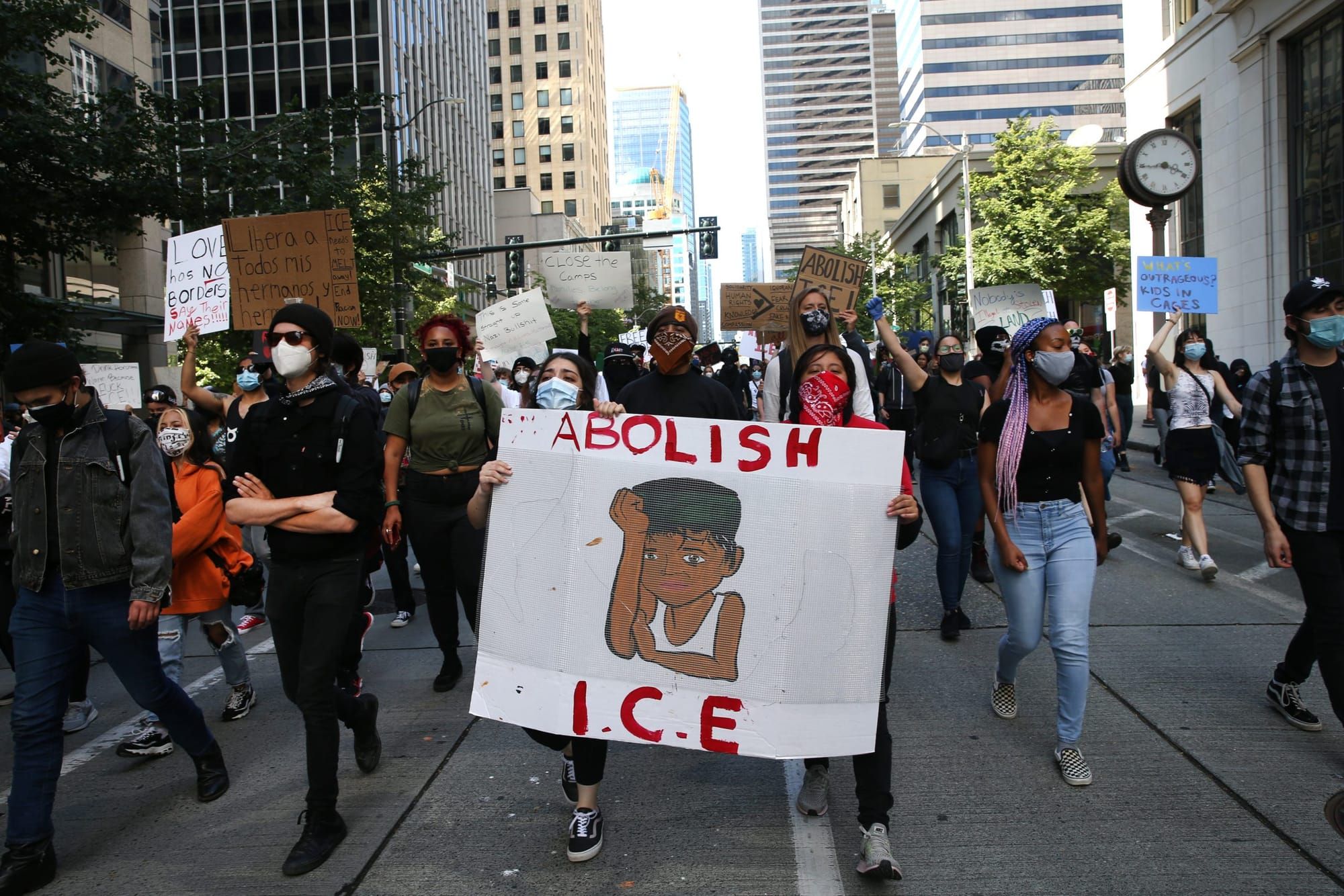 color photograph of an urban outdoor protest. people wear face masks and walk in the middle of a shaded street. two women sta