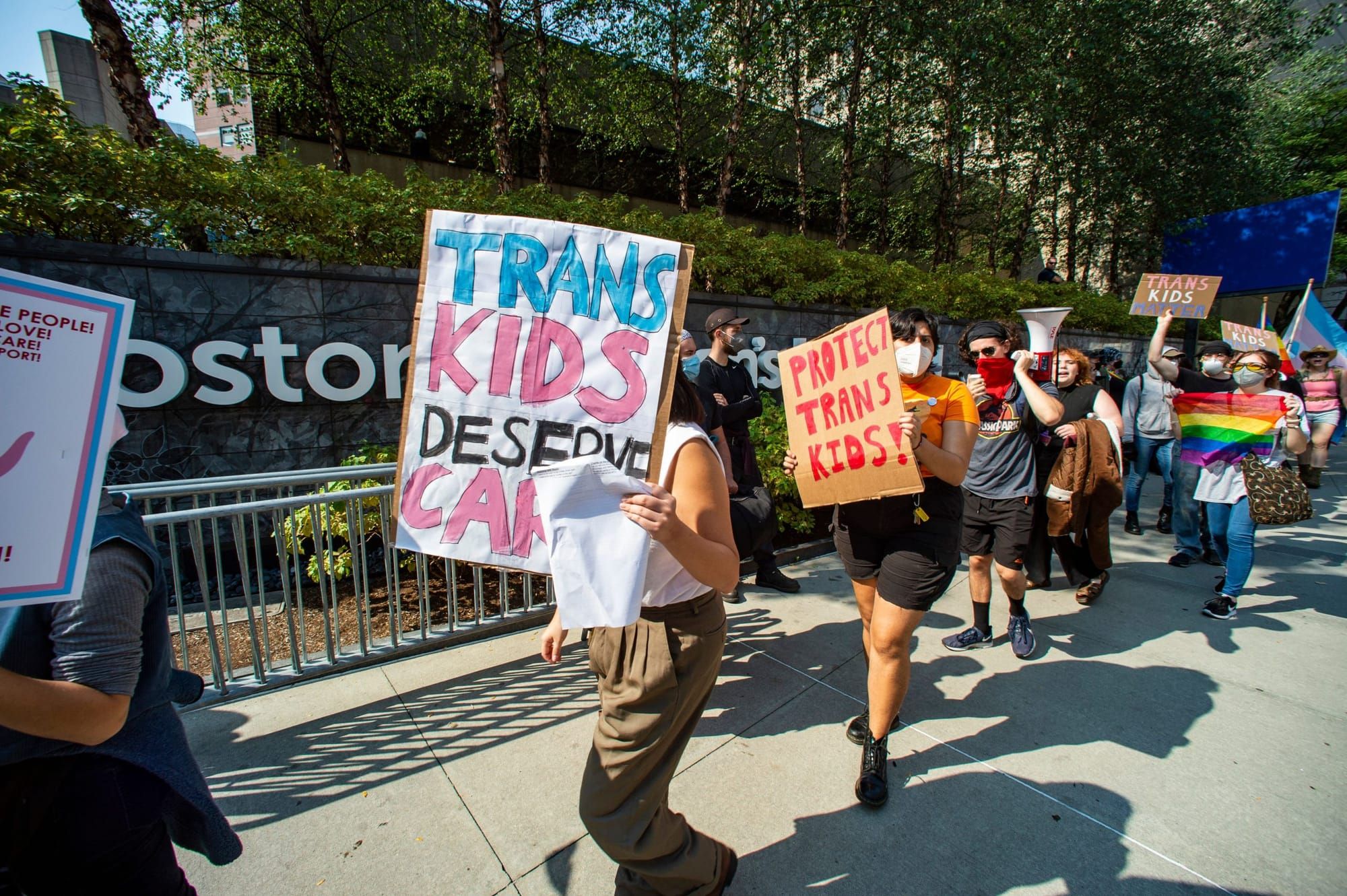 color photograph of an outdoor protest where protesters are walking in a single-file line carrying signs in support of trans