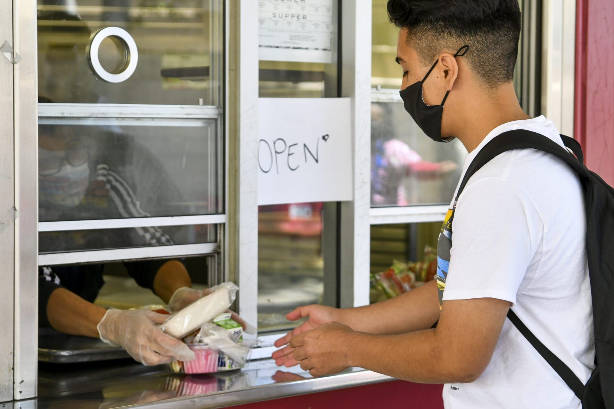 color photograph of a high school boy with an undercut fade wearing a white t-shirt and a black backpack received food from a