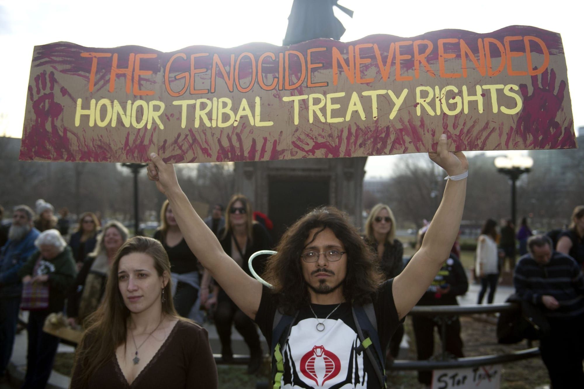 color photograph of an outdoor protest in front of an elevated statue where a man with shoulder-length hair holds up a cardbo