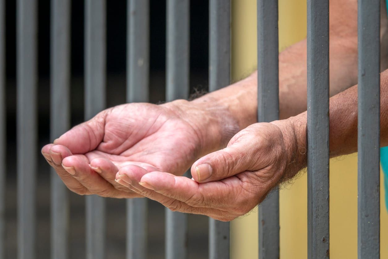 color stock photo of an elderly person's hands cupped upwards sticking through prison bars