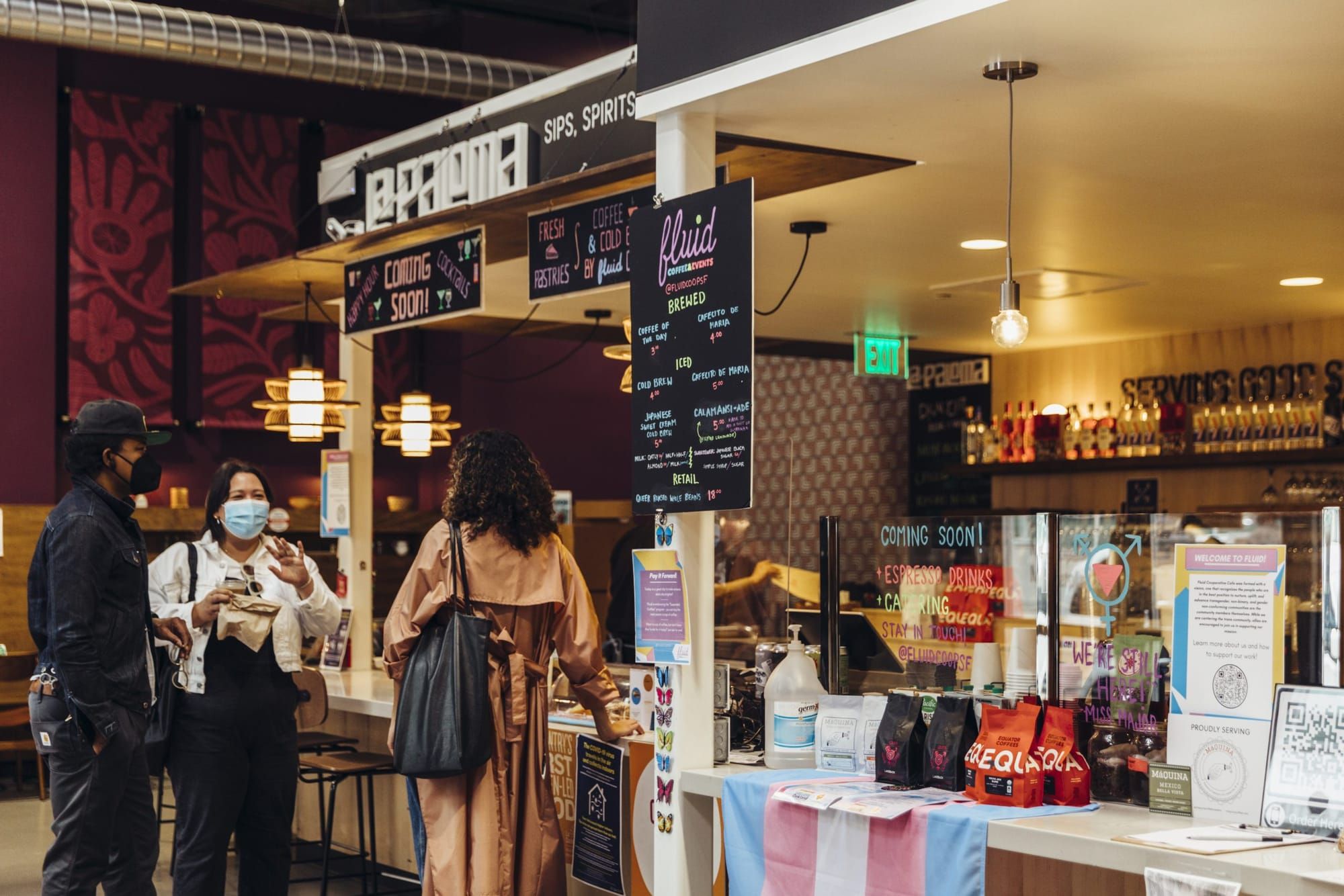 color photograph of an indoor marketplace stall that sells coffee and espresso. a trans flag hangs over the counter, and thre