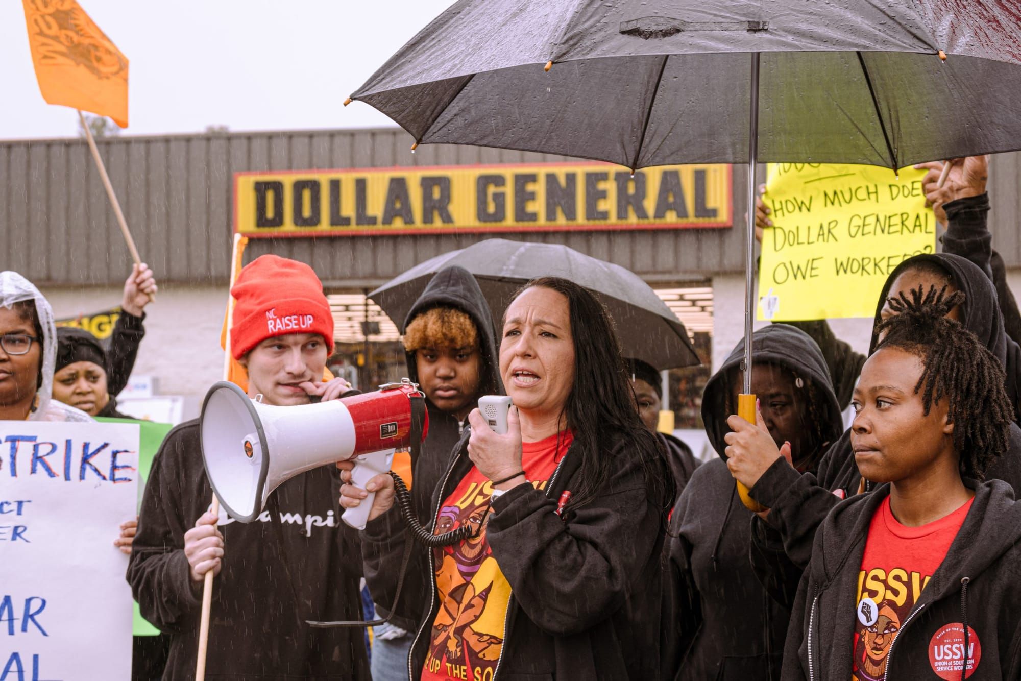 color photograph of a latina woman holding a red and white bullhorn microphone in front of a small crowd of people holding si