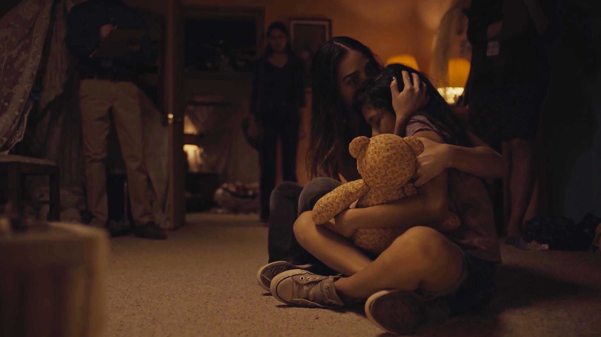 color movie still of a Latinx woman sitting cross-legged on the floor of a house. she is holding her young daughter in her la