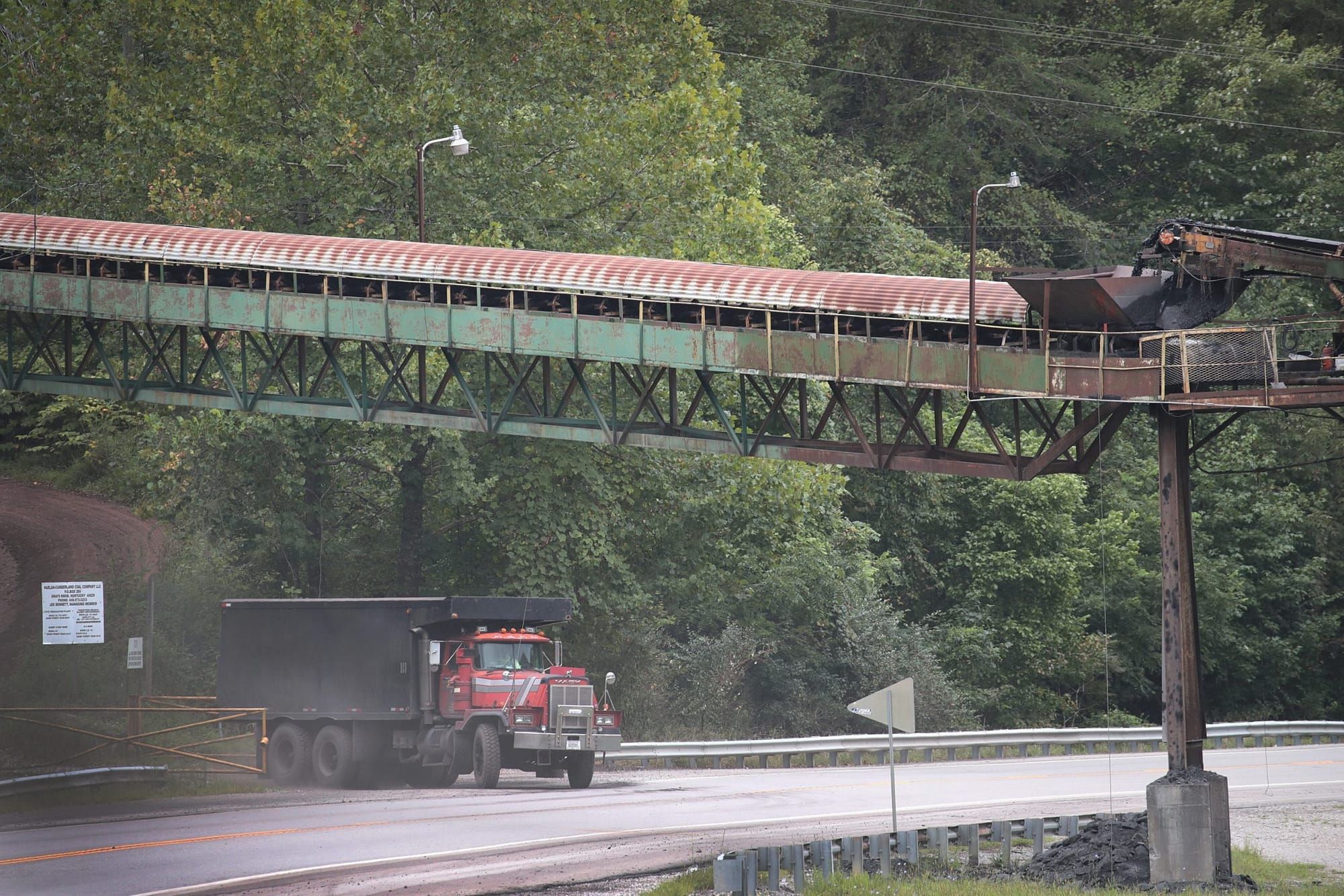 color photograph of a truck leaving a forested area onto a paved street. a metal overpass stands above the truck in the foreg