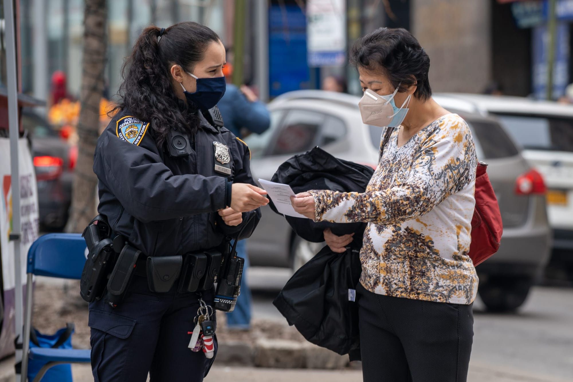 color photograph of an NYPD officer wearing a long-sleeved jacket handing a pamphlet to and older Asian woman outside on a Ne