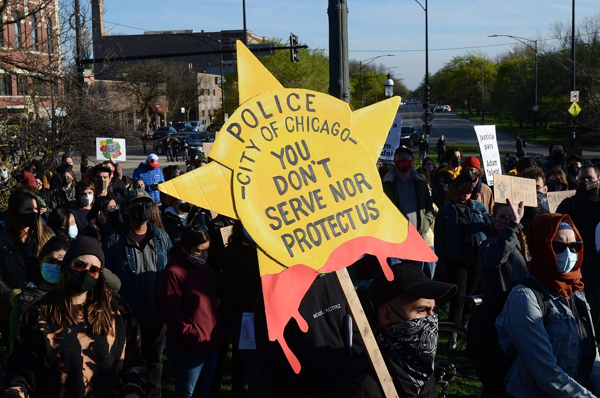color photograph of an outdoor protest against the police. in the foreground is a yellow sign in the shape of a police badge