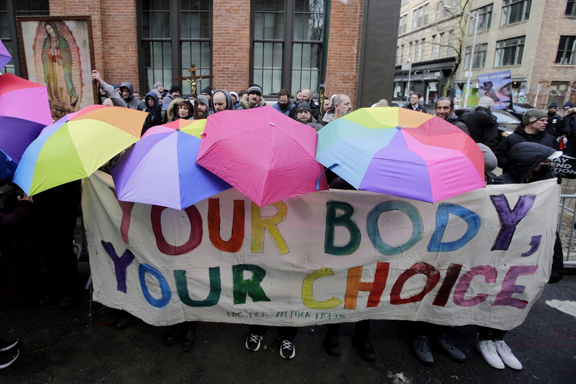 color image of an outdoor protest where people stand obscured under rainbow umbrellas and carry a large banner in rainbow let