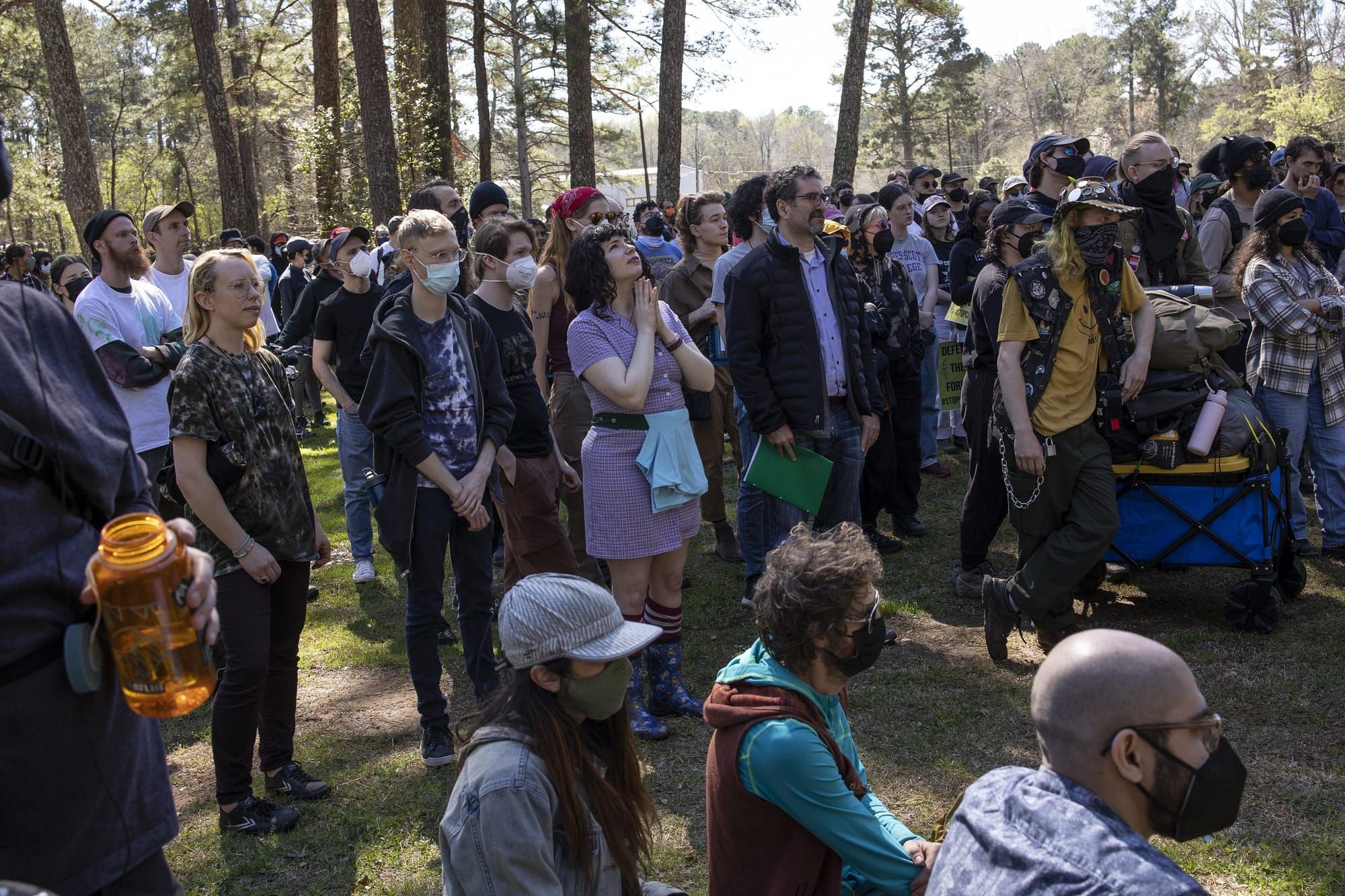 color photograph of about fifty people standing and sitting outside in a forest clearing. many wear face masks, t-shirts or f