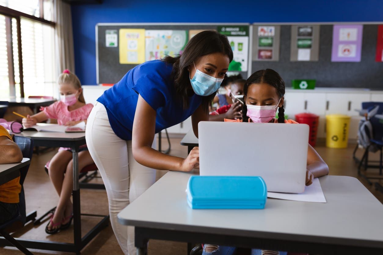color stock photo of an African american female teacher wearing face mask teaching a girl to use laptop at elementary school