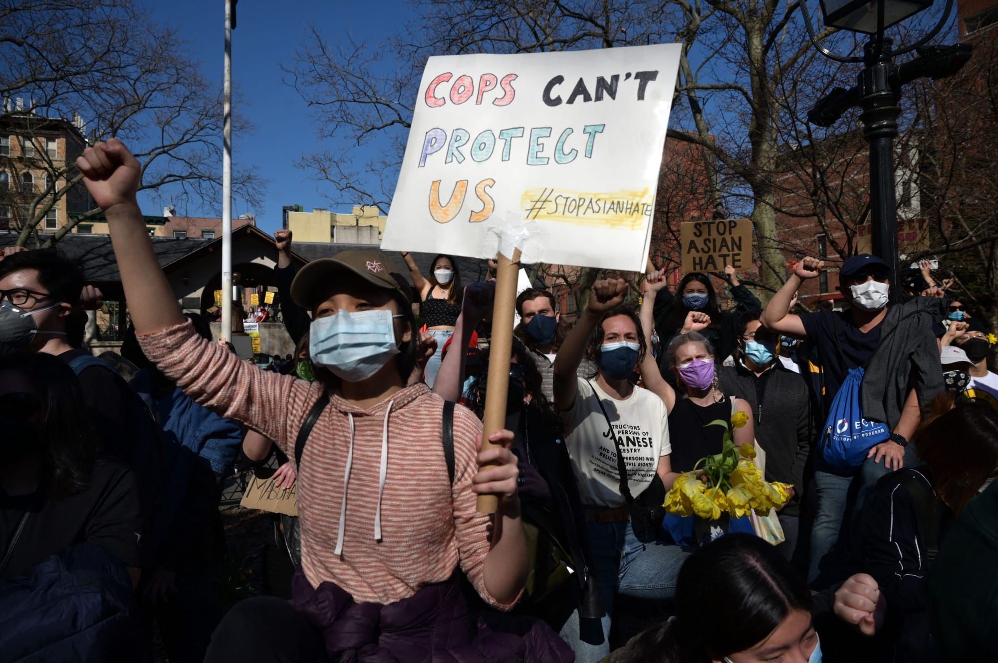 color photograph of an outdoor protest. in the foreground, an asian femme person wearing a dusty pink hoodie and a medical ma
