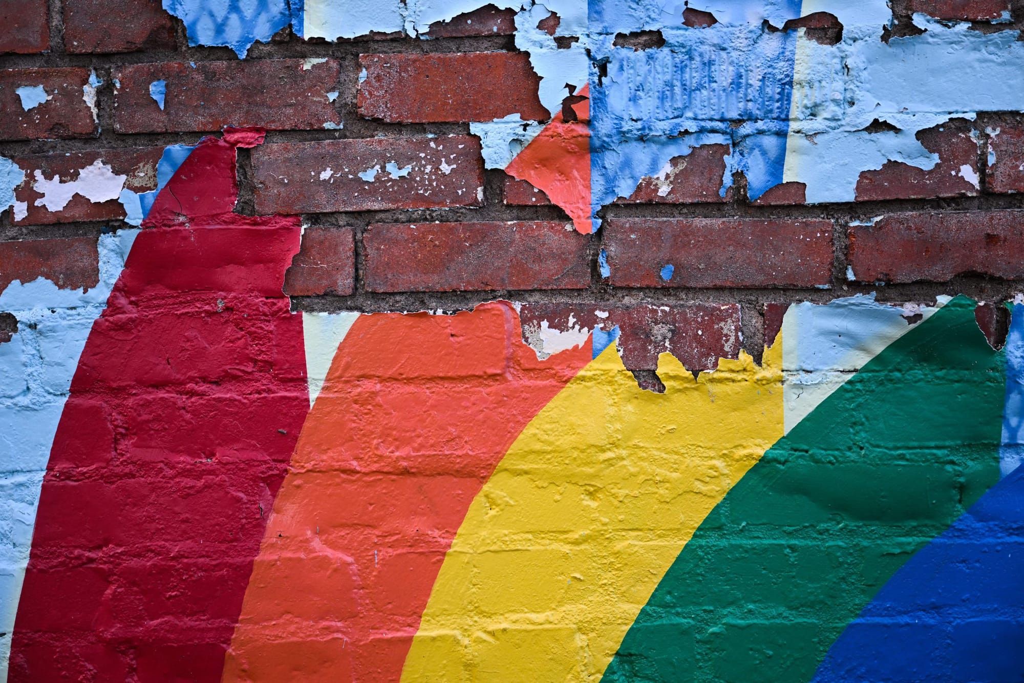 color photograph of a rainbow poster partially torn through the middle, exposing brick wall behind it