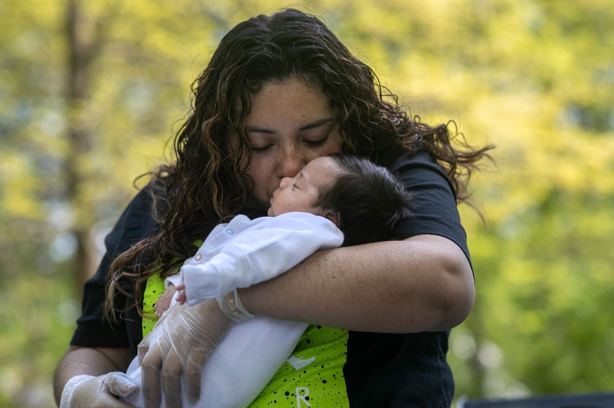 color photograph of a latinx mother with chest-length curly hair holding a 6-week-old infant in her arms and kissing his chee