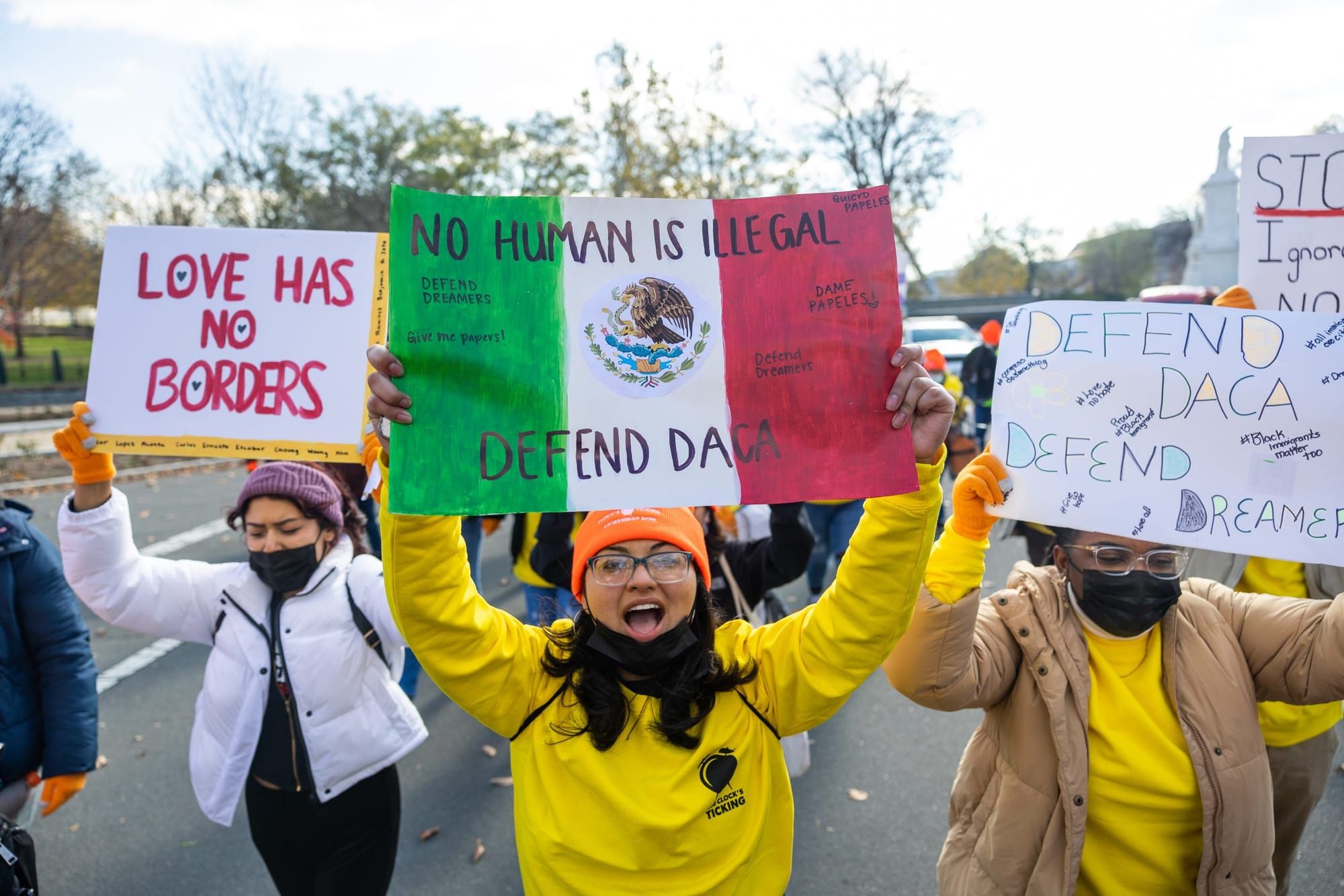 color photograph of an outdoor protest. people hold up signs in support of DACA. a person in a white puffy jacket on the left