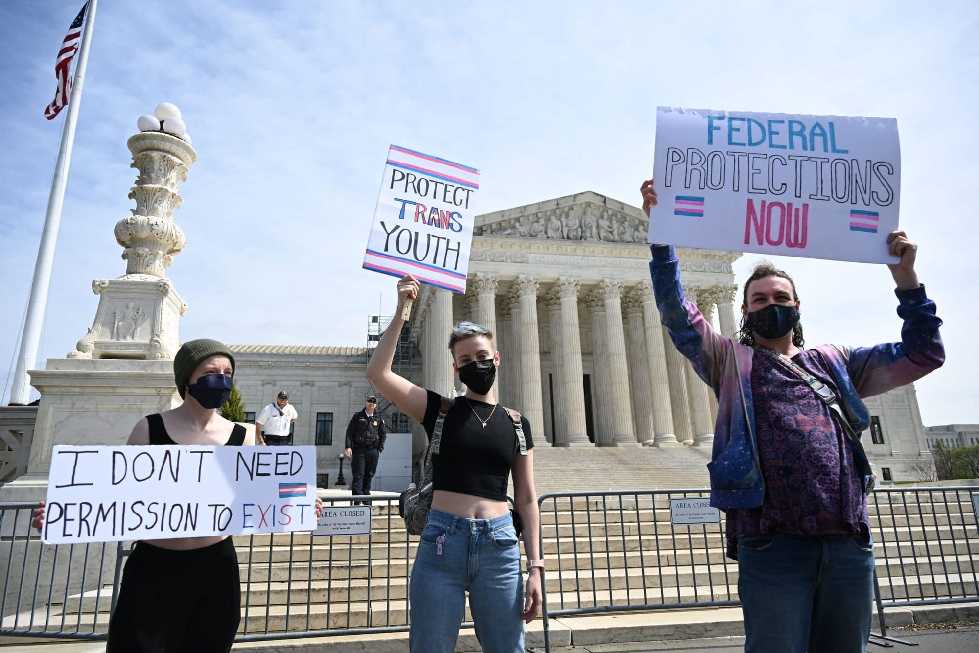 color photograph of three protesters outside the U.S. Supreme Court building. they hold white posters with trans flags and pi