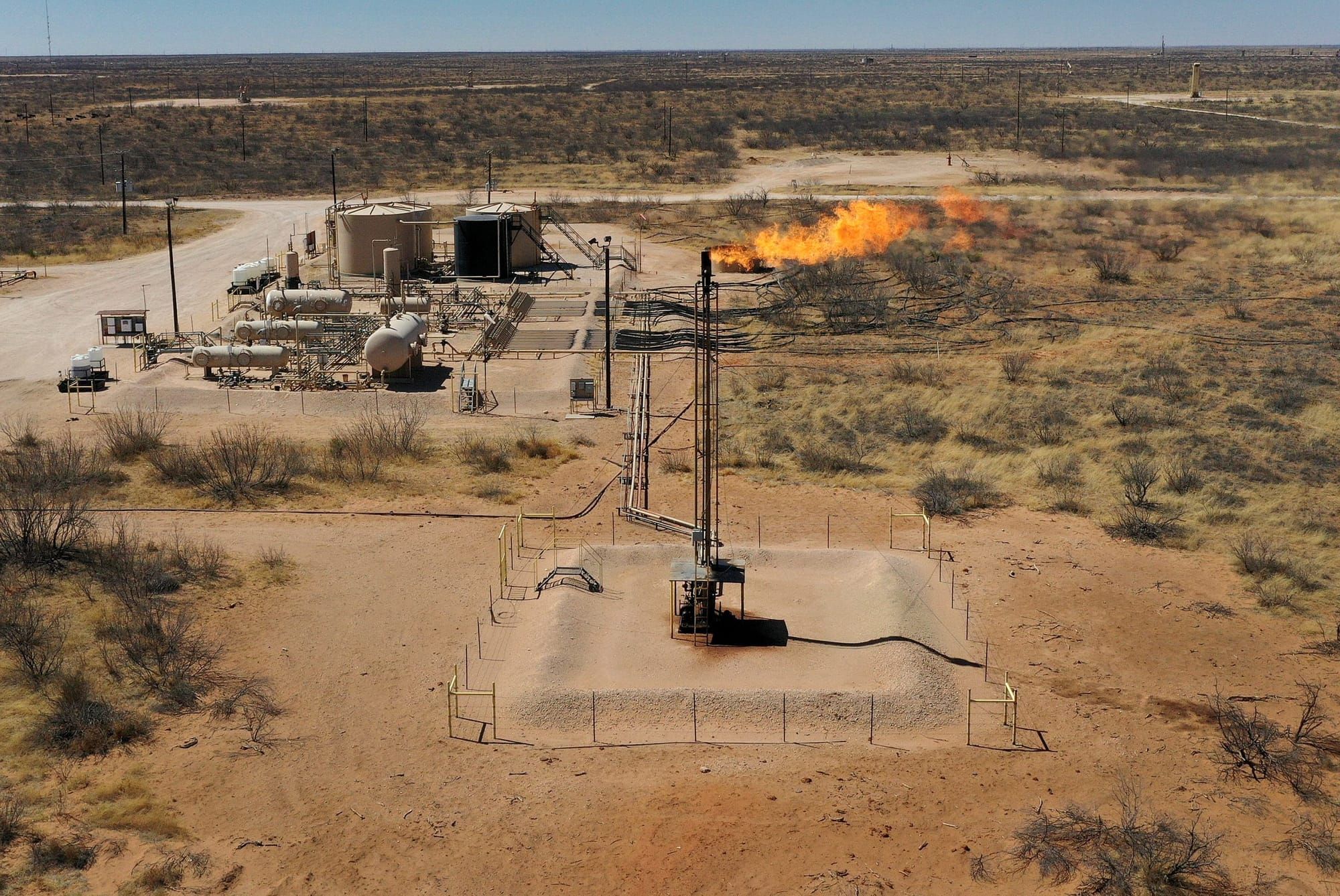 color photograph of a flaring natural gas rig surrounded by dusty sand and sparse shrubs