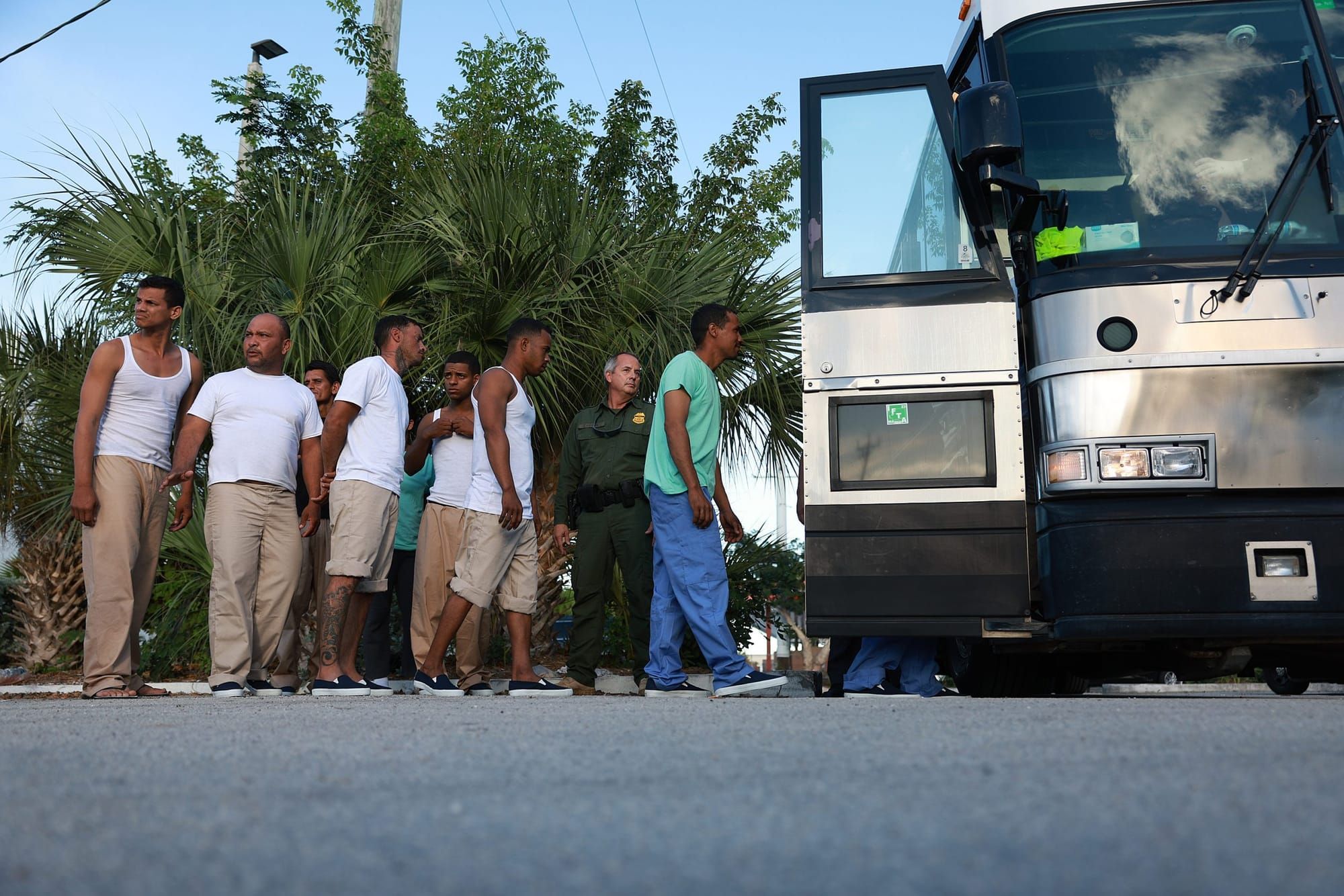 outdoor color photograph of about six Latinx men in white t-shirts and khaki pants standing in line waiting to board a bus