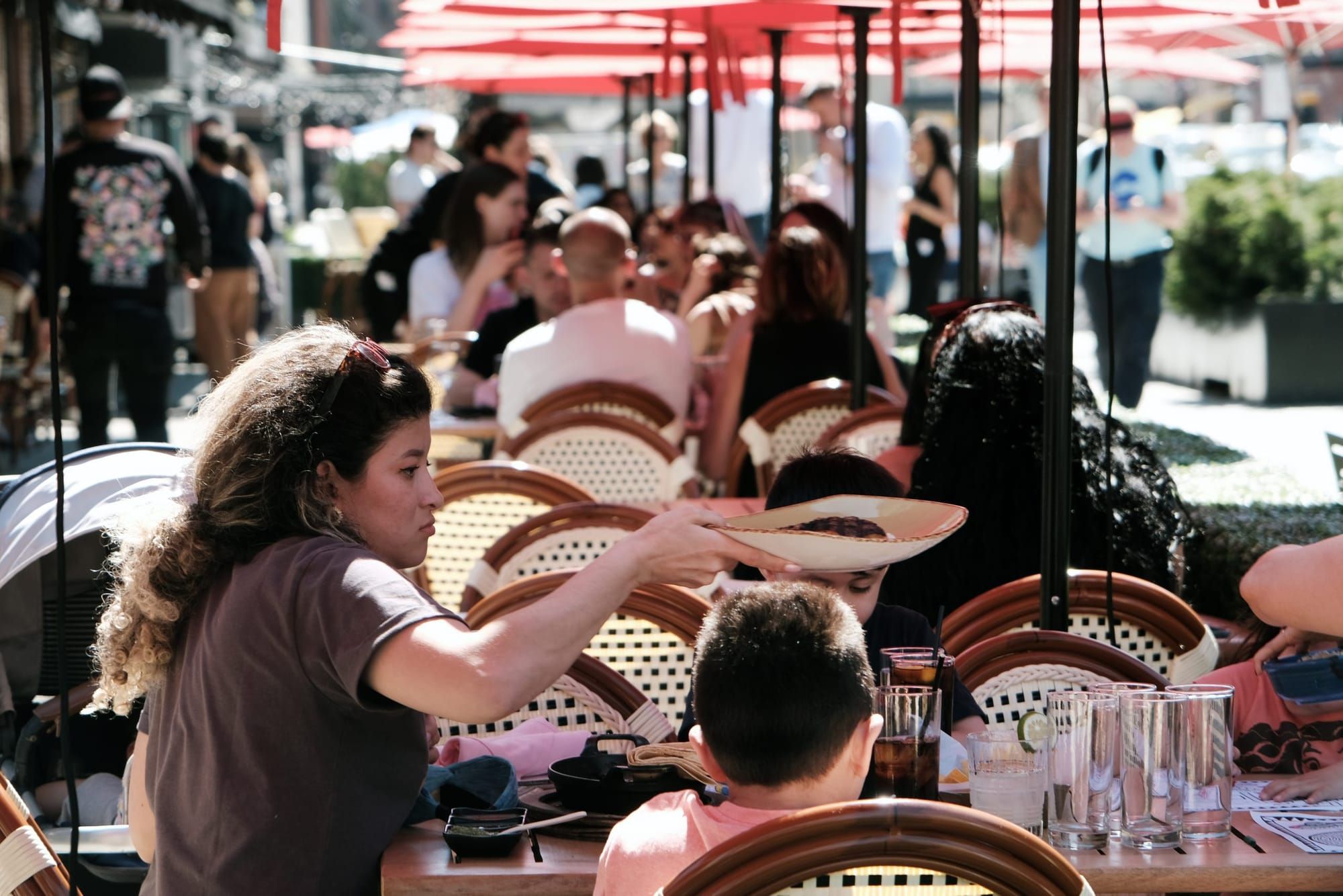color photograph of a Latinx woman holding a plate above a seated person's head at an outdoor restaurant with red overhead um