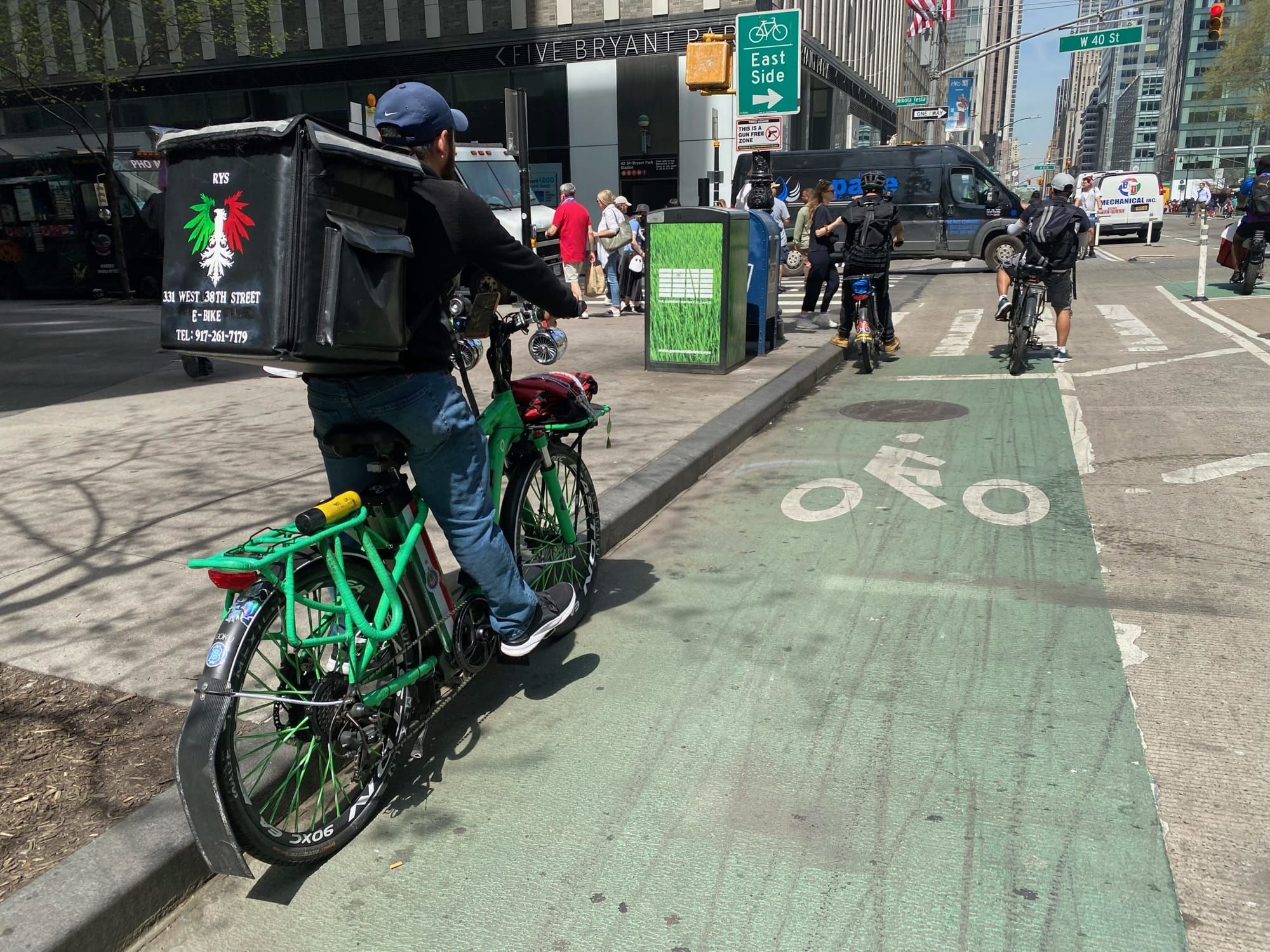 color photograph of the back of a delivery person riding an e-bike. They have a large cube pack on their back with an eagle t