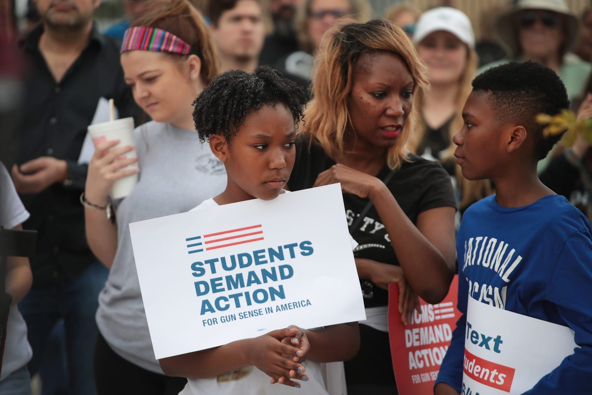 color photograph of an outdoor student protest for gun control. a Black student stands in the foreground and holds a large wh