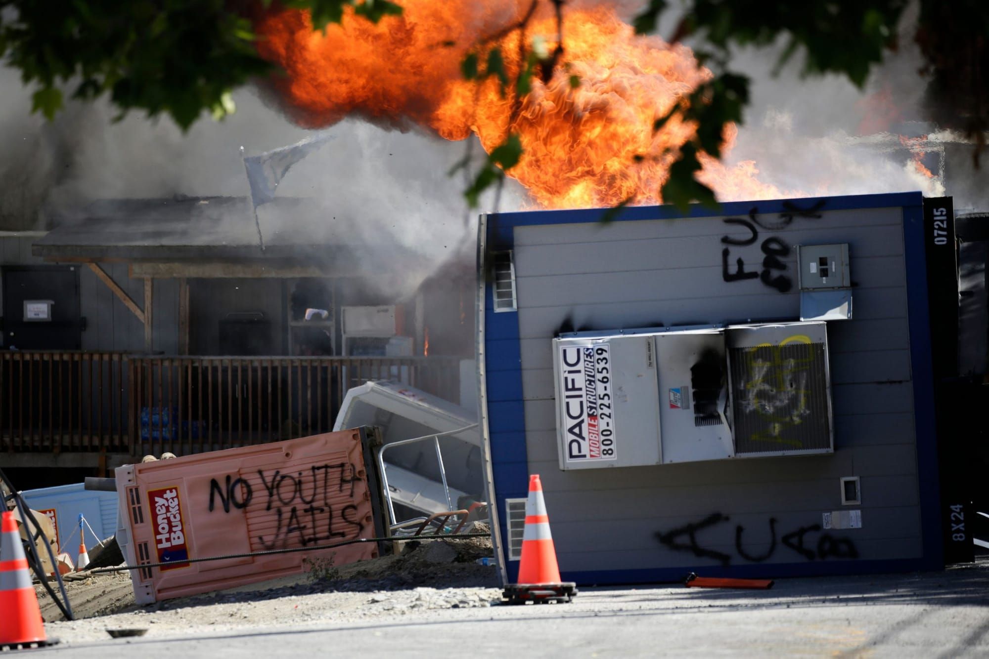 color photograph of a trailer on fire. to the left is an overturned port-a-potty with black graffiti spray painted on it that