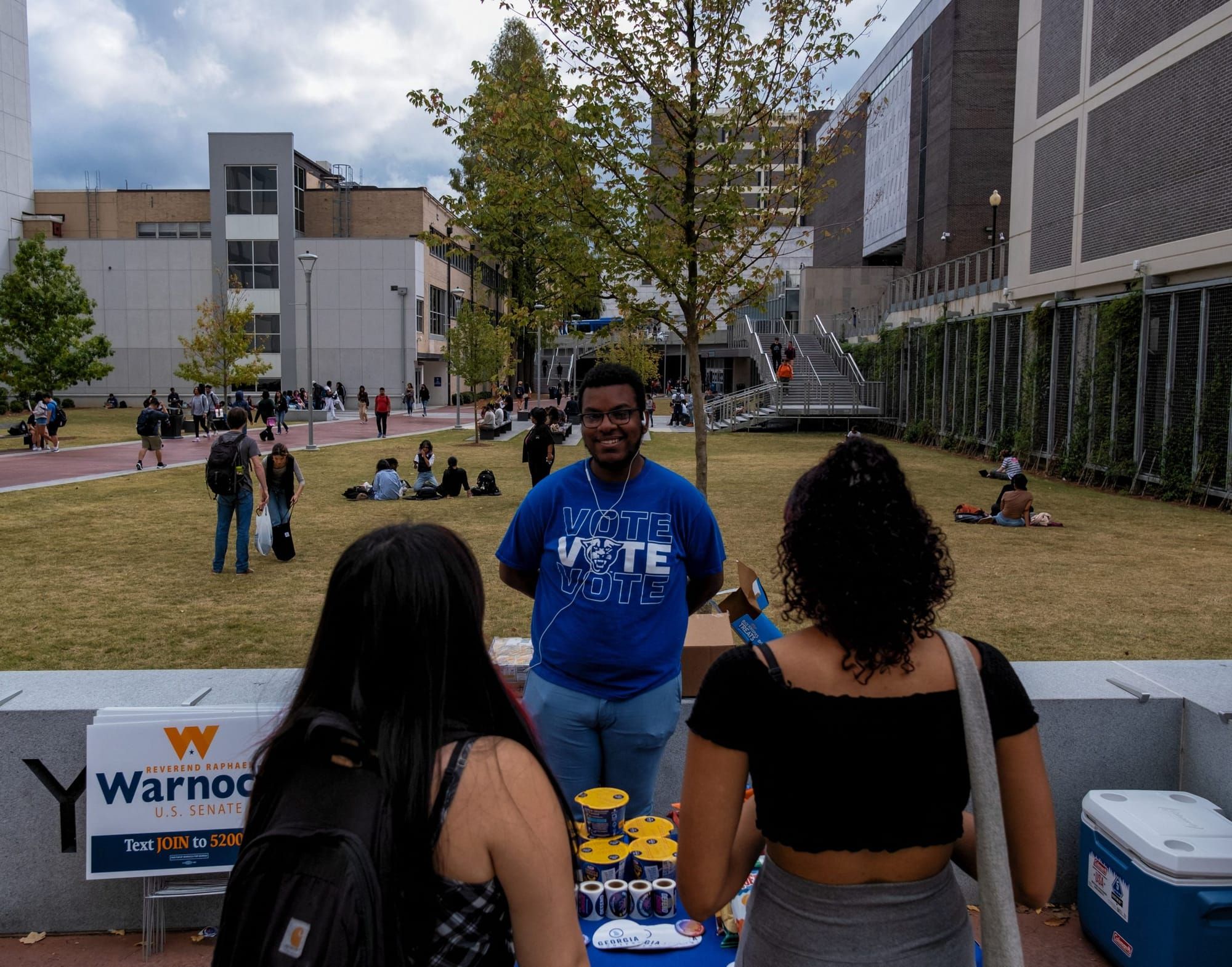 color photograph of a Black young man in a dark blue shirt with white text that reads "VOTE" on it faces the camera. Two peop