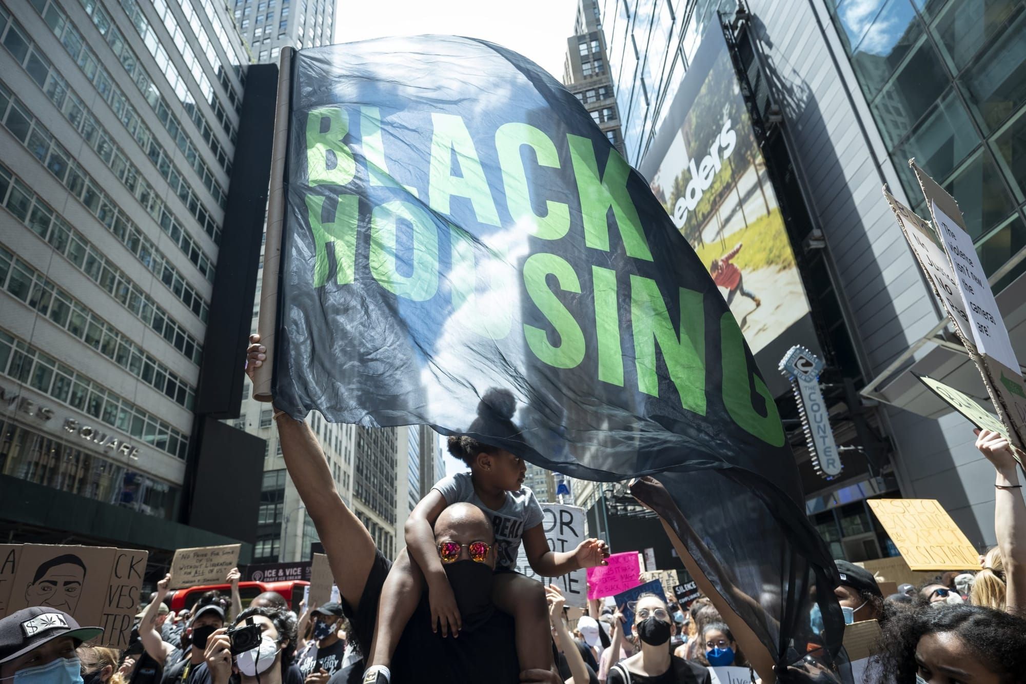 color photograph of an outdoor protest. a black man carries a young child on his shoulders and waves a black flag with large