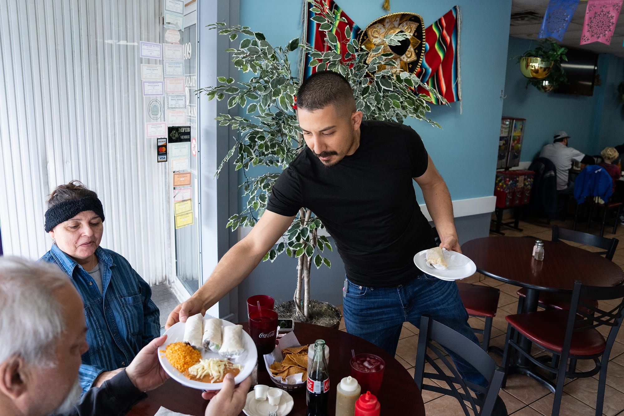 color photograph of a Latinx man with buzzed hair wearing a black t-shirt and blue jeans setting down plates at a round woode