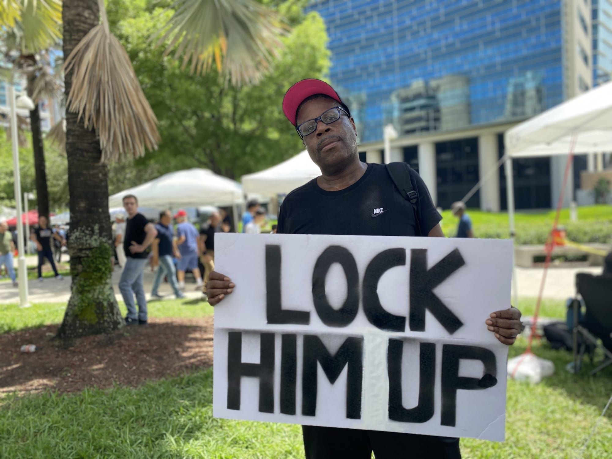 color photograph of a Black man standing outside in a green area between commercial buildings. He wears a black shirt and a b