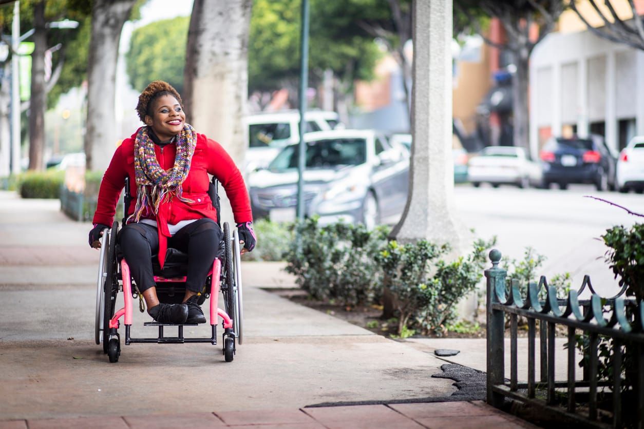 color photograph of a Black woman wearing a bright red zip-up and black pants sits in a wheelchair and travels up a sidewalk