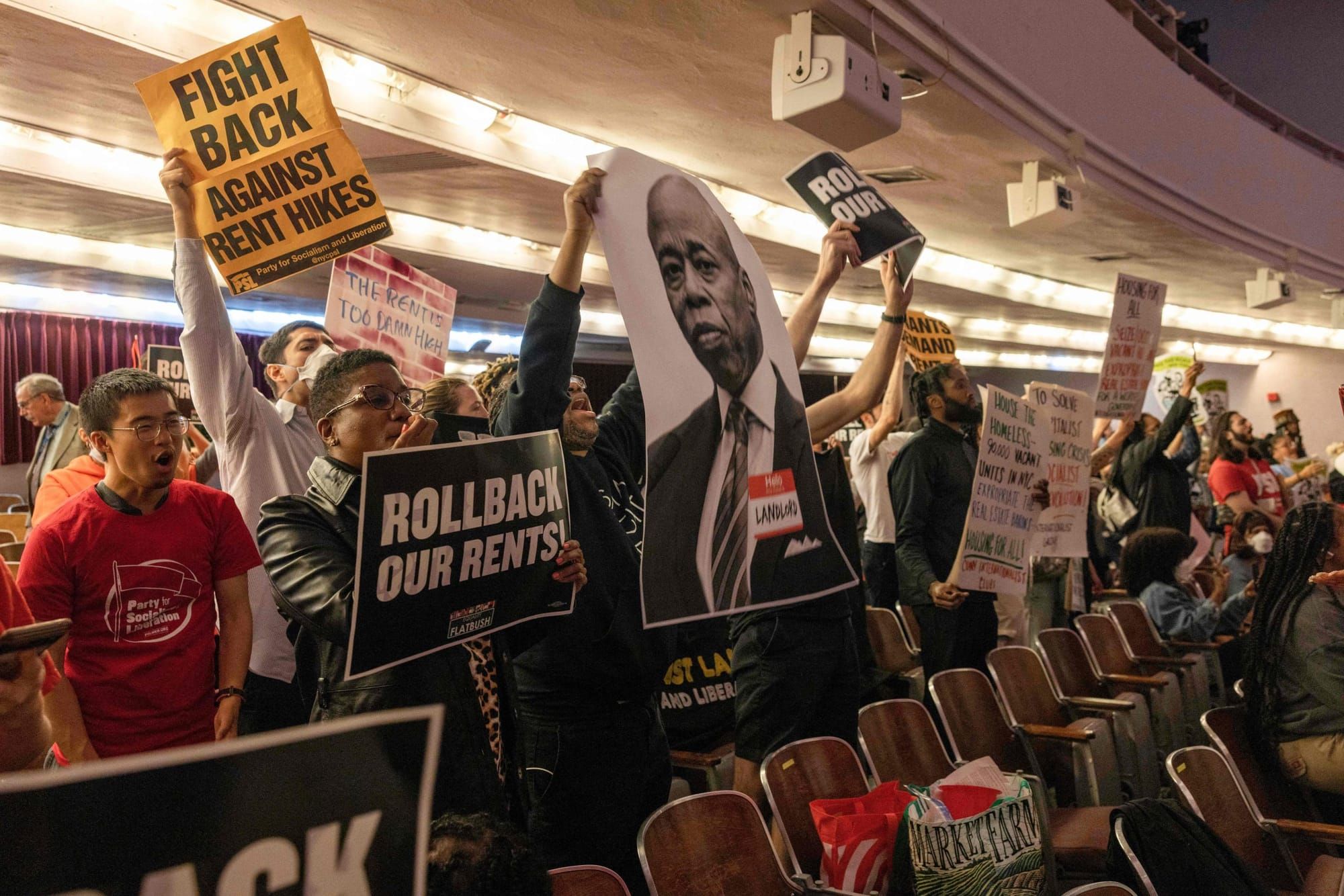 color photograph of an indoor protest for tenants' rights. people hold up posters of new york city mayor eric adam along with