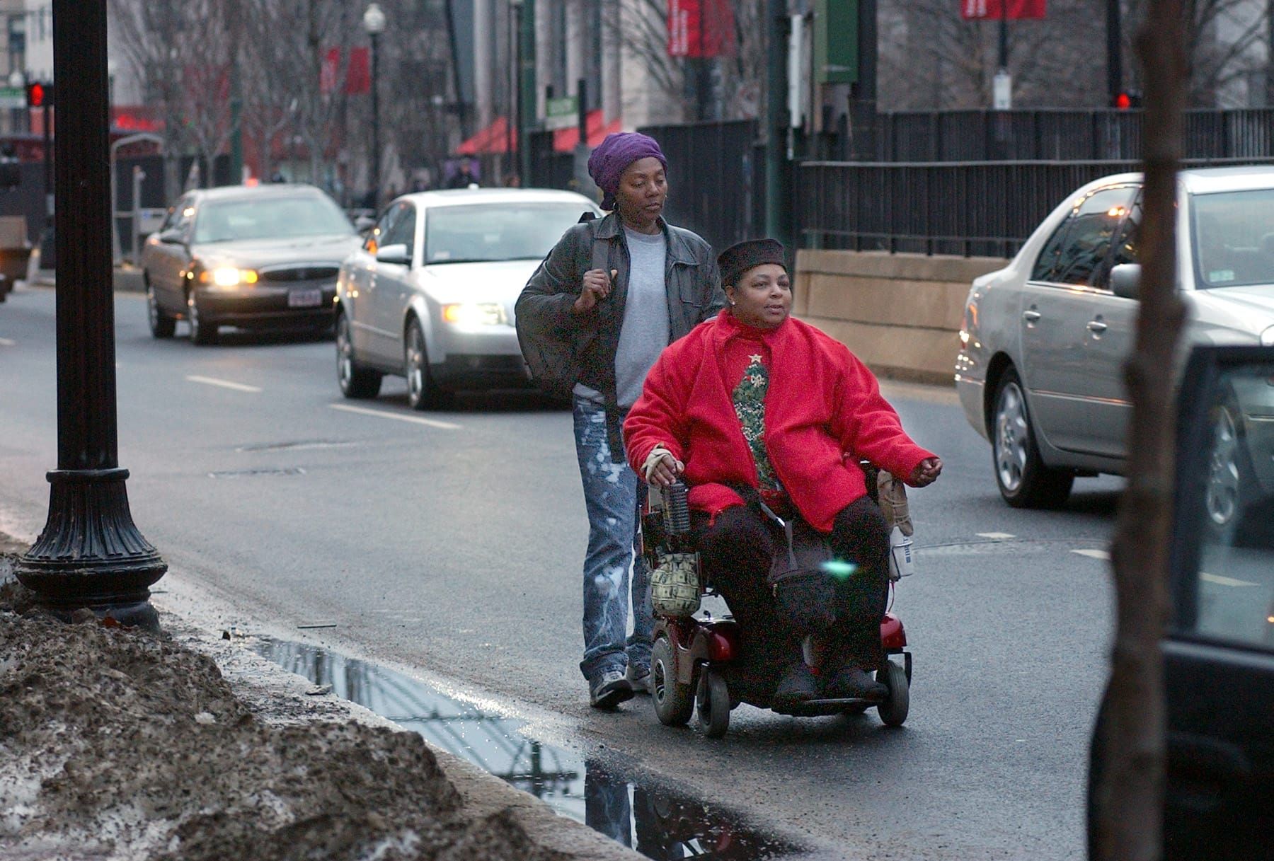 color photograph of a Black woman in a wheelchair traveling on a paved street. another Black woman walks behind her, and cars