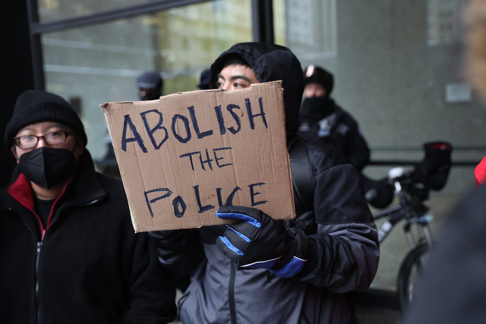 color photograph of a protester wearing a black winter coat and holding a cardboard sign with handwritten text that reads "ab