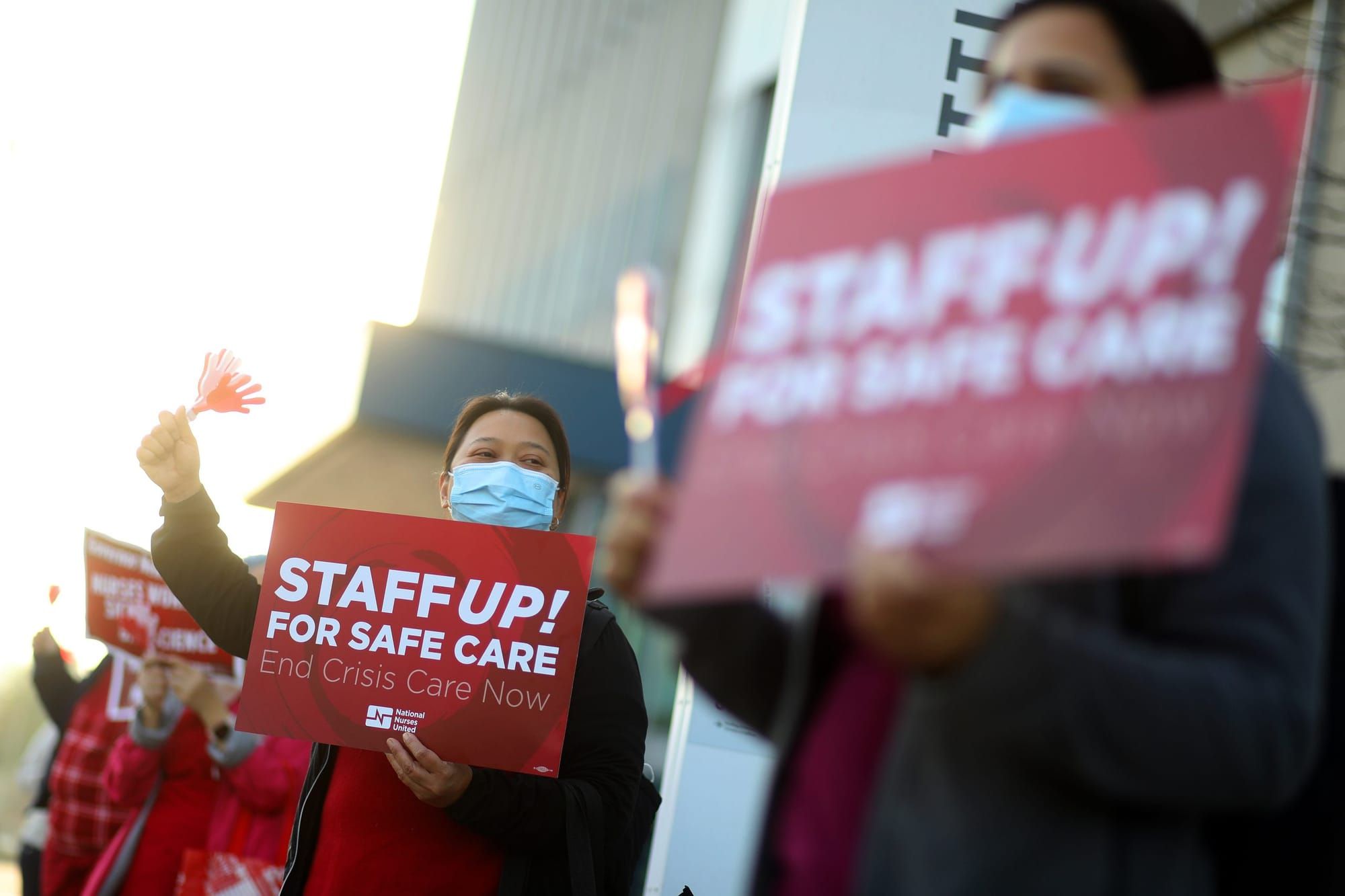 color photograph of nurses wearing masks and holding up red signs with printed white serif-font text that reads "staff up! fo